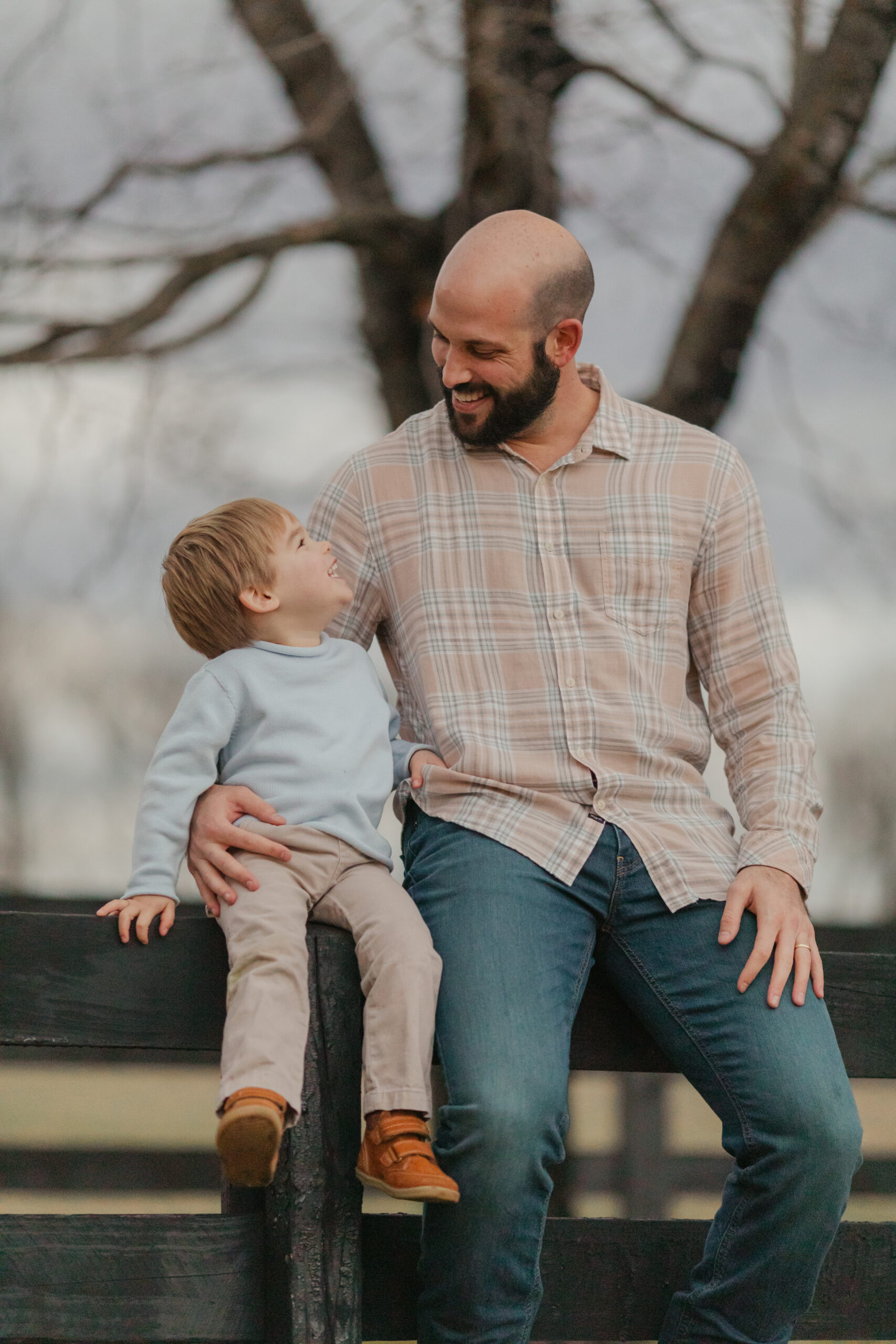 a dad and his 2 year old son sit on the top of a black fence, smiling at each other as they pose for family photos at a park in Nashville, Tn. 