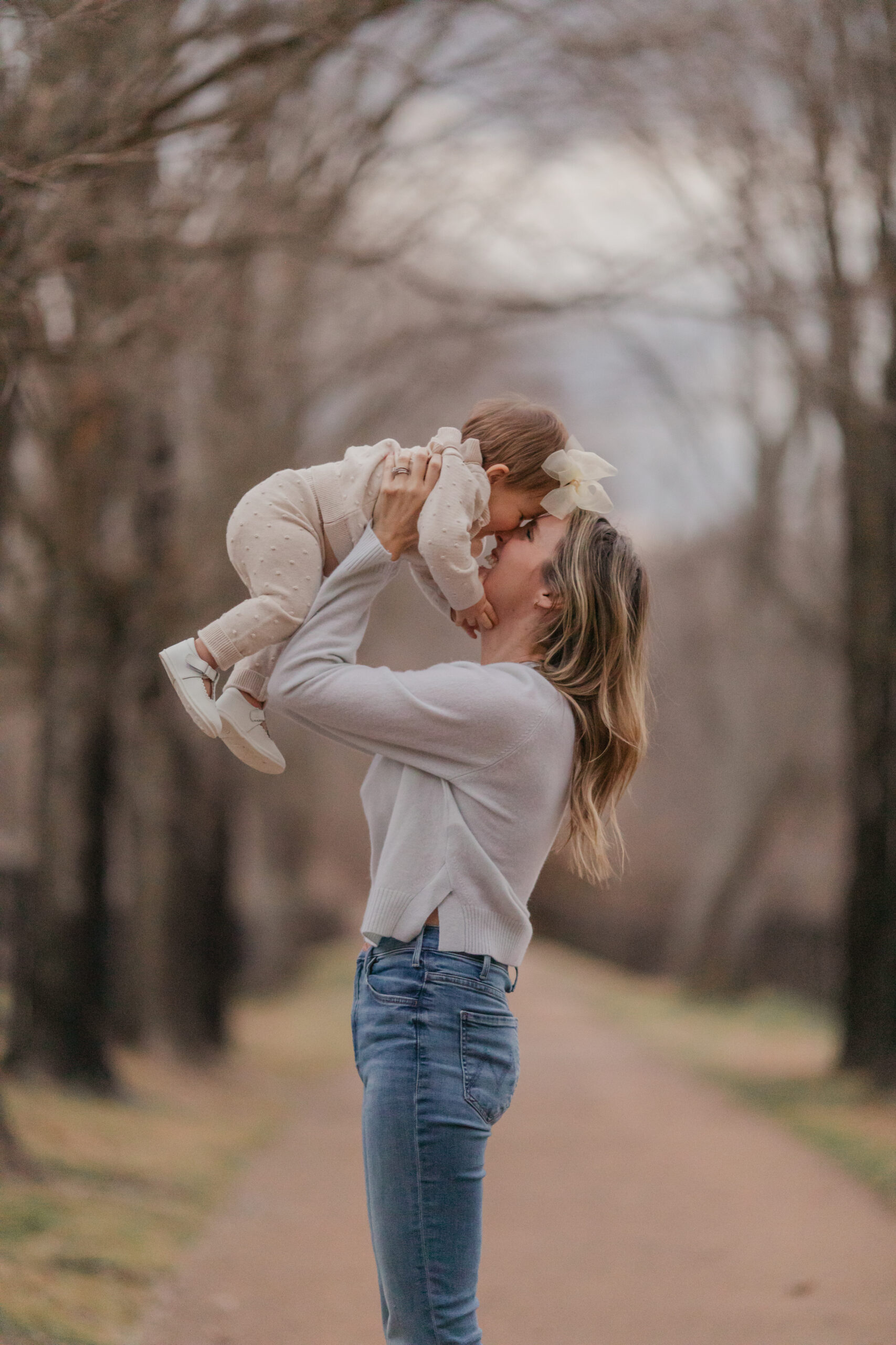 mom and baby girl play together on a winter day in a driveway lined with trees that create a tunnel overhead.