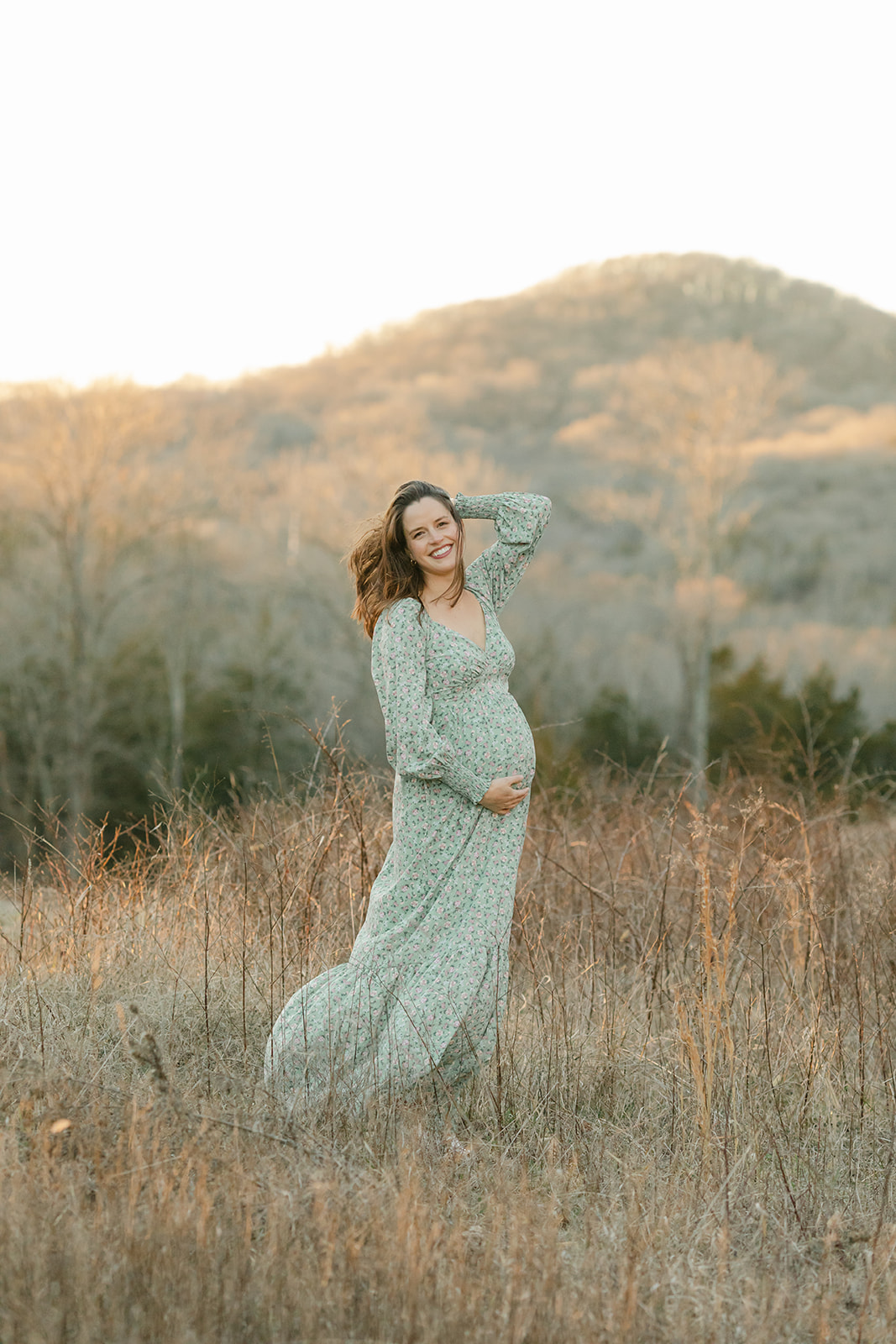 A pregnant woman wearing a blue floral dress stands in tall dried winter grass, with rolling hills in the distance behind her. She is laughing as the wind blows through her hair.