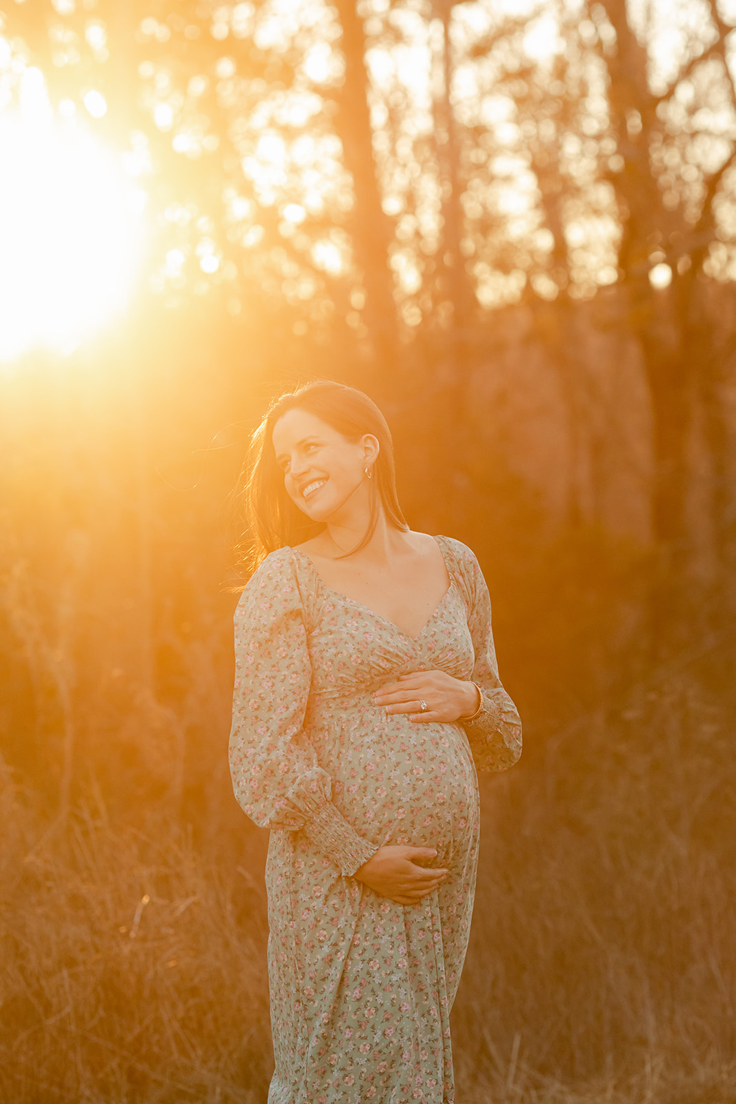 a pregnant woman in a blue dress stands next to the forrest edge as golden sunset light dances on the winter tree branches, in brentwood tennessee.