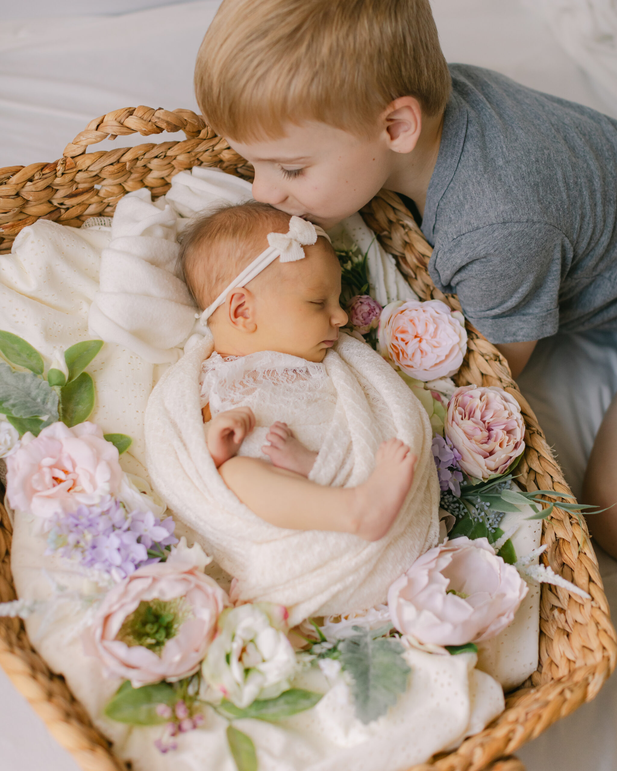 big brother kissing sleepy newborn baby sister on the head while she's in basket with flowers