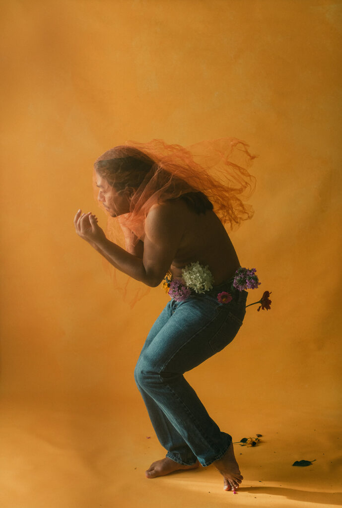 queer man poses shirtless with flowers coming out of the tops of his jeans while standing in front of a yellow backdrop