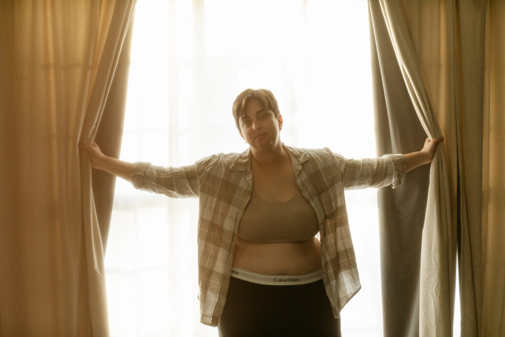 gender non-conforming queer woman poses by opening the curtains of a window while standing in her underwear