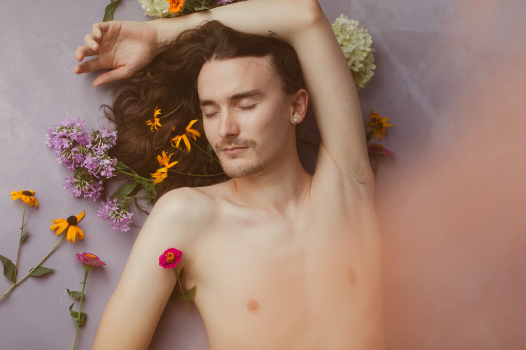 topless man lays with his eyes closed surrounded by flowers 